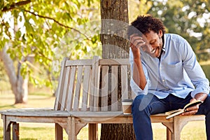 Mature Man Relaxing Sitting On Park Bench Under Tree Reading Book With Takeaway Coffee