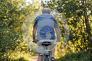 Mature man is riding bicycle in forest path with the bike to the far end