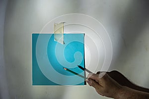 Mature man`s hand points to a piece of paper with a pencil. Empty blue square piece of paper fixed to the wall. An abstract light