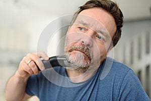 Mature man is shaving off his beard with electric razor at home during quarantine. Handsome bearded man trimming his beard with