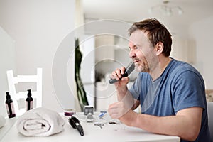 Mature man is shaving off his beard with electric razor at home during quarantine. Handsome bearded man trimming his beard with