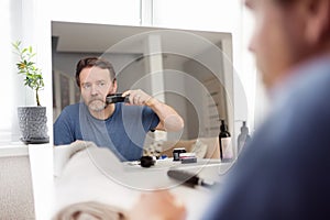 Mature man is shaving off his beard with electric razor at home during quarantine. Handsome bearded man trimming his beard with