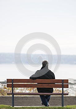 Mature man sitting alone overlooking water on park bench in Autumn.