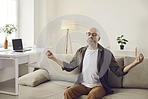 Mature man sitting on couch at home, meditating, trying to relax and manage stress