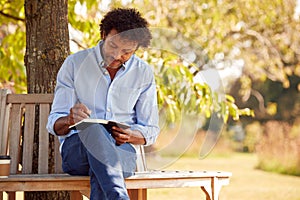 Mature Man Sitting On Park Bench Under Tree Writing In Notebook