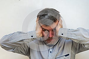 A mature man in a state of psychological stress in front of a light wall. A male with a short haircut and graying hair covers his
