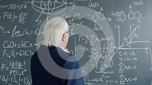 Mature man in suit walking to chalkboard wall with formulas looking at equations