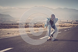 Mature man in sunglasses with hands back riding his longboard on country road with mountains in background. Man skating or