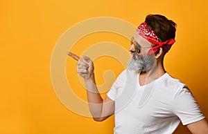 Mature man in sunglasses, red bandana, white t-shirt. Smiling, pointing at something by forefinger, posing on orange background