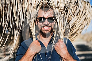 A mature man with sunglasses standing outdoors on beach, straw umbrella on his head.