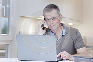 Mature man talking on smart phone in front of a laptop at home