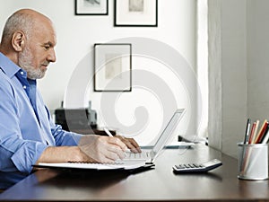 Mature Man Using Laptop And Writing In Notepad