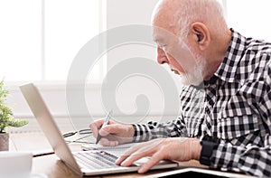 Mature man using laptop and writing in notepad