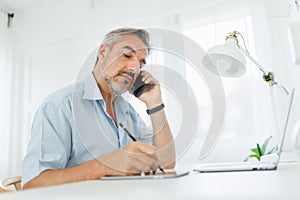 Mature man using mobile phone while writing something on digital tablet