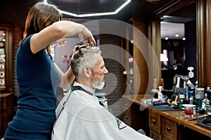 Mature man visiting hairstylist. Side view of professional barber girl doing haircut for elegant middle aged man sitting