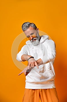Mature man in white hoodie, pants and sunglasses, bracelets. Showing cool sign by his hands, posing on orange background. Close up
