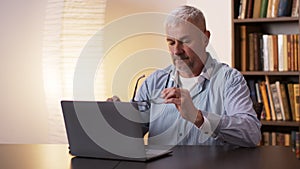Mature man wiping and putting on glasses and working with laptop computer at home office