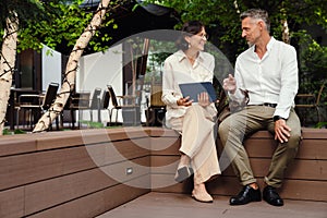 Mature man and woman discussing business details and using tablet while sitting outdoors