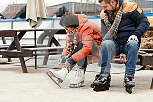 Mature man and woman getting ready to skate on ice rink outdoors. Woman tying laces on white skates. Active weekends