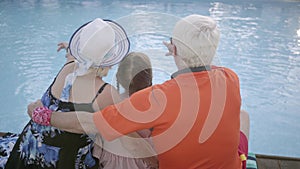 Mature man, woman and little girl sitting on the edge of the pool, back view. Grandmother, grandfather, and grandchild