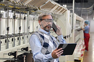 Mature Manager Looking At Digital Tablet Screen And Using Phone In Textile Factory