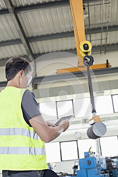 Mature manual worker with digital tablet operating crane to lift steel in industry