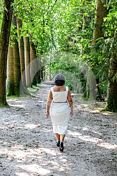 Mature Mexican woman in a white dress walking away from the camera on a dirt path between trees in the forest