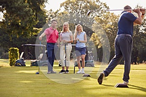 Mature And Mid Adult Couples Standing By Golf Buggy Watching Man Hit Tee Shot