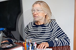 Mature middle aged in glasses using laptop typing email working at home office, lady searching information on internet
