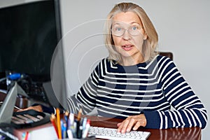 Mature middle aged in glasses using laptop typing email working at home office, lady searching information on internet