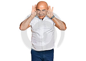 Mature middle east man with mustache wearing casual white shirt trying to hear both hands on ear gesture, curious for gossip