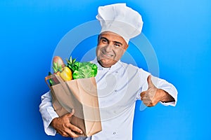 Mature middle east man professional chef holding bag of groceries smiling happy and positive, thumb up doing excellent and