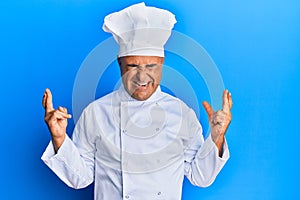 Mature middle east man wearing professional cook uniform and hat gesturing finger crossed smiling with hope and eyes closed