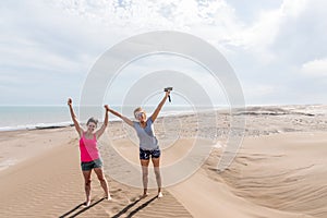 Mature mother and adult daughter with their arms raised, on a large dune with the beach in the background.