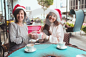 Mature mother and her young daughter sit together in cafe or restaurant. Women sit togeher wearing santa hats and