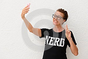 Mature nerd woman wearing big eyeglasses and standing against white background outdoors while taking selfie picture and