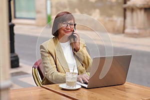 Mature office employee woman talks on smartphone with smile about upcoming business trip while sitting on terrace in