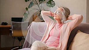 Mature old 60s woman, older middle aged female resting on couch taking deep breath of fresh air holding hands behind