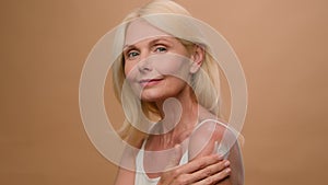 Mature old Caucasian woman in beige studio apply moisturizing body lotion sunscreen cream on shoulder middle aged lady