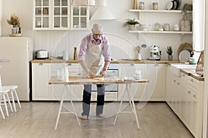 Mature older baker man in apron rolling dough on table