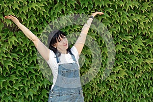 A mature pretty middle-aged woman with dark long hair stands against the background of a green fence and holds her hands up