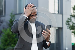 Mature senior african american boss reading bad news on phone, businessman in business suit outside office building