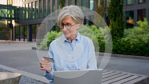 Mature senior Caucasian businesswoman business woman pay bills with bank credit card and laptop outdoors old female