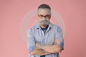 Mature serious man with crossed arms wear blue shirt isolated on pink background.