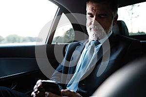 Mature smiling businessman using smartphone while sitting on back seat of business car. Happy senior man reading email.