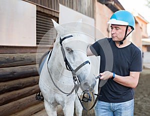 Mature smiling man farmer in helmet standing with white horse at stable