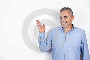 Mature smiling man with grey hair wearing blue shirt standing on isolated white background holds his hand up and to the