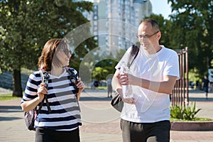 Mature smiling man and woman walking in city park talking drinking water from bottle
