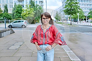 Mature smiling woman in embroidered shirt looking at camera, on street of modern city