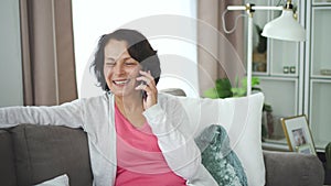 Mature smiling woman talking phone sitting on couch in home interior.